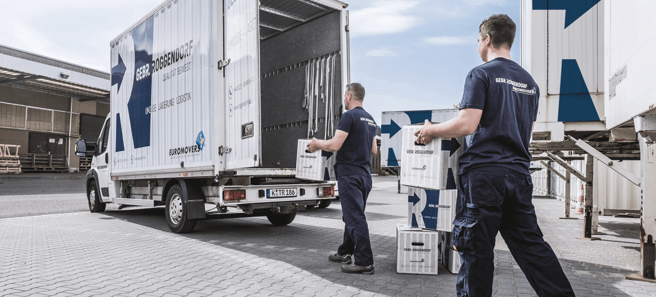 Zwei Umzugshelfer laden Kartons mit der Aufschrift "AKTEN" in einen weißen Transporter an einem Gewerbestandort.